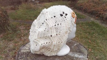 Cremeweiße gehäkelte Beanie mit Relief- und Lochmuster und zwei Holzknöpfen auf einer Büste im Freien.