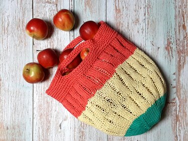 Gestrickte Einkaufstasche in Koralle, Gelb und Türkis mit roten Äpfeln auf hellem Holz