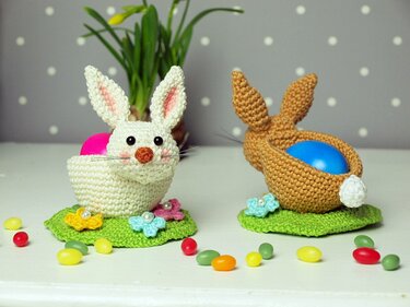 Zwei gehäkelte Osterhasen-Eierhalter, weißer Hase mit rosa Ei und brauner Hase mit blauem Ei auf grünen Häkelunterlagen, umgeben von bunten Süßigkeiten und Häkelblumen.