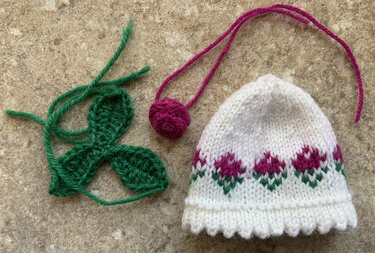 Weißer gestrickter Eierwärmer mit rosa Blumenbordüre, grünem Blattmuster, gehäkelte Blätter und pinke Bommelkordel