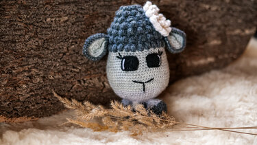 Gehäkeltes Amigurumi-Schaf in Grau mit schwarzen Augen und weißer Blüte, auf weichem Untergrund vor Baumrinde