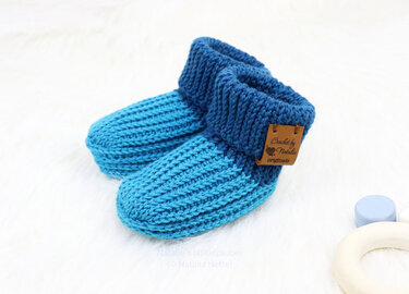 Two-tone blue crocheted baby booties with ribbed cuffs on a white background with a wooden toy