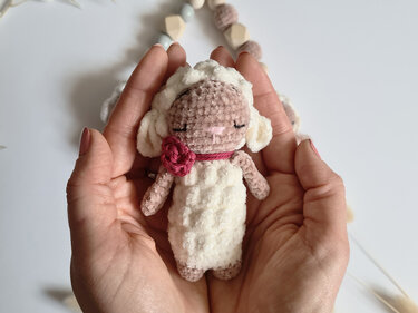 Gehäkeltes Mini-Lämmchen mit weißem flauschigem Körper und rosa Blüte, in Händen gehalten