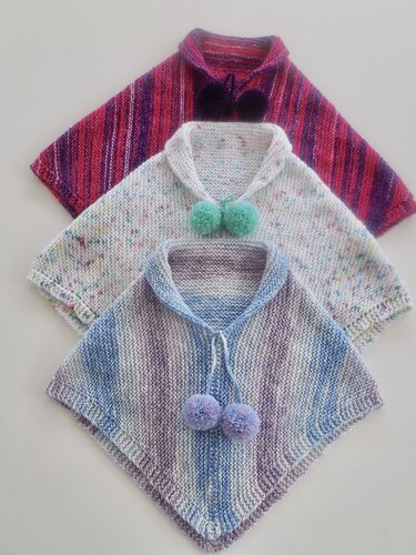 Drei gestrickte Kinderponchos (rosa‑lila, weiß gesprenkelt, blau‑lila) mit Pompons auf hellem Hintergrund