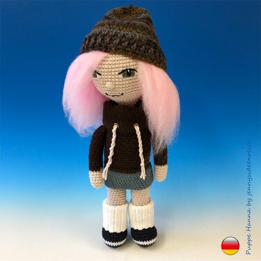 Gehäkelte Amigurumi‑Puppe mit rosa Haaren, brauner Mütze, Hoodie und weißen Stiefeln vor blauem Hintergrund