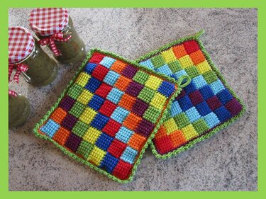 Zwei bunte handgehäkelte Topflappen mit kariertem Muster und grüner Umrandung auf einer Arbeitsfläche; im Hintergrund drei Gläser mit karierten Deckeln.