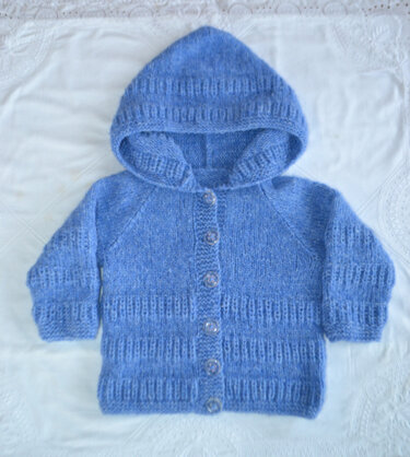 Gestrickte hellblaue Kapuzenjacke für Babys mit Knopfleiste, liegend auf hellem Stoff