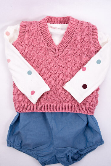 Rosa gestrickte ärmellose Babyweste mit VAusschnitt ber weißem Langarmshirt mit bunten Punkten und blauer Pumphose