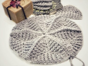 Runder grauer gehäkelter Untersetzer mit Reliefmuster, daneben Seife und weitere Untersetzer.