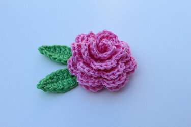 Gehäkelte pinke Rose mit zwei grünen Blättern auf hellem Hintergrund