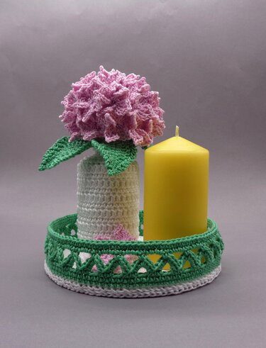 Rosa gehäkelte Blume in weißem Häkelgefäß neben gelber Kerze auf grün-weißem Häkeltablett