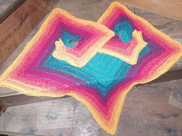 Zwei gehäkelte quadratische Ponchos mit Farbverlauf in Türkis, Pink und Gelb auf Holz