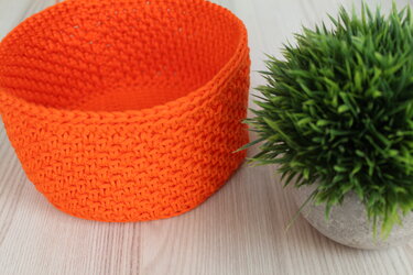 Gehäkelter runder Korb in Orange neben einer kleinen grünen Topfpflanze auf hellem Holztisch.