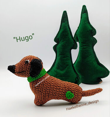 Gehäkelter brauner Dackel Amigurumi mit grünem Halsband und Herz vor zwei grünen Filzbäumen