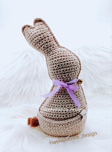 Gehäkelter beiger Osterhase mit lila Schleife und Bommel-Schwanz, sitzend auf weißem Fell