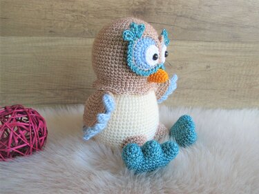 Beige-cremefarbene Amigurumi-Eule mit hellblauen Akzenten und orangefarbenem Schnabel auf weißem Fell.