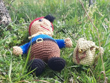 Handgehäkelte Amigurumi-Figur in blauem Oberteil neben kleinem Häkelbeutel mit Glöckchen im Gras