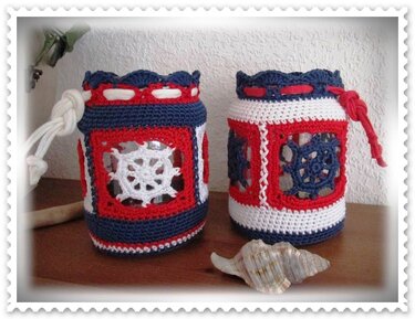 Zwei maritime gehäkelte Bezüge in Rot, Weiß und Blau um Kerzengläser mit Steuerradmotiven