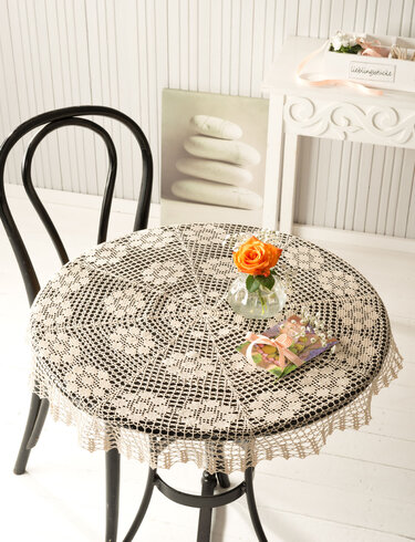 Rundes gehäkeltes Spitzen-Deckchen auf kleinem Tisch mit Glasvase und orangefarbener Rose