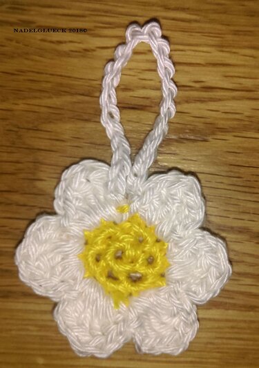 Gehäkelte weiße Margeritenblume mit gelbem Zentrum und Aufhängeschlaufe auf Holzuntergrund