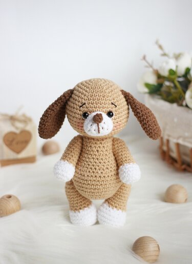 Beige crocheted amigurumi dog with brown floppy ears and white paws standing on a neutral surface