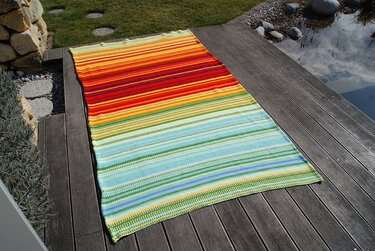 Bunte gehäkelte Decke mit warmen Rot- und Gelbtönen oben und kühlen Blau-Grün-Tönen unten auf einer Terrasse neben einem Teich