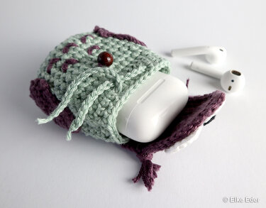 Mint and purple crocheted owl pouch with a white earbud charging case partly inside and two white earbuds nearby.
