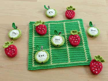 Green crocheted tic-tac-toe board with red strawberry and round white fruit tokens on a wooden table