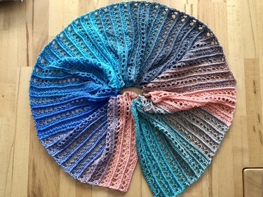 Halbrunder gehäkelter Schal mit Farbverlauf in Türkis, Blau, Grau und Rosa, aufgefächert auf hellem Holzboden