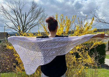 Person von hinten hält ein weißes gehäkeltes Dreieckstuch mit Rautenmuster vor gelb blühenden Sträuchern