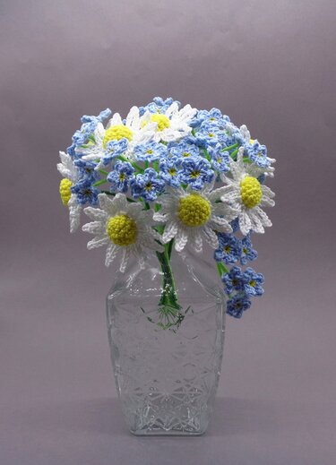 Gehäkelter Blumenstrauß mit weißen Margeriten und blauen Vergissmeinnicht in einer transparenten Glasvase vor neutralem Hintergrund.