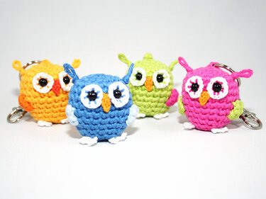 Four colorful crocheted amigurumi owl keychains in blue, pink, green and orange on a white background
