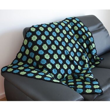 Gehäkelte Decke aus blauen und grünen Granny-Squares mit schwarzer Umrandung auf grauem Sofa