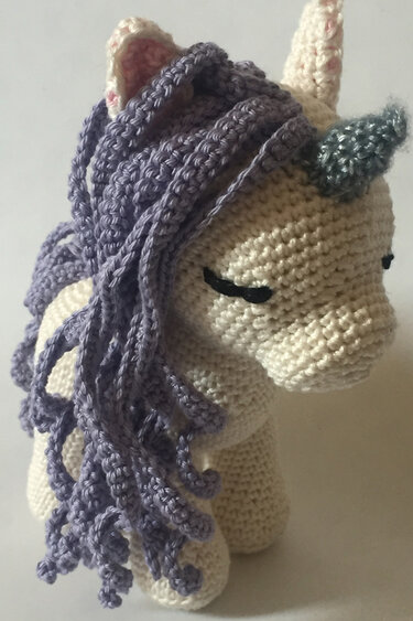 Close-up of a crocheted amigurumi unicorn with a cream body, purple curly mane, pink inner ears, and a bluish-grey horn.