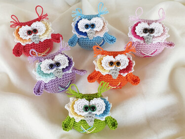 Sechs farbenfrohe gehäkelte Amigurumi-Eulen mit auffälligen Augen auf hellem Stoff.