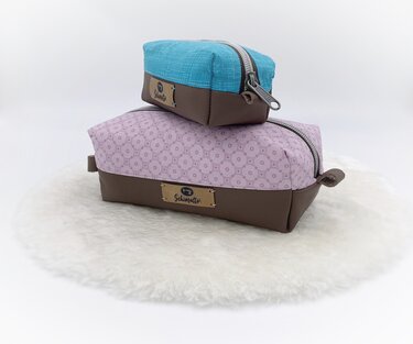 Zwei rechteckige Kosmetiktaschen, eine rosa gemustert und eine türkis, beide mit braunem Boden in Lederoptik und Reißverschluss, auf hellem Untergrund