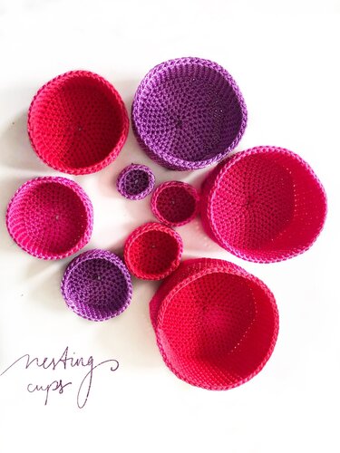Neun gehäkelte Stapelbecher in verschiedenen Größen in Rosa-, Pink- und Lilatönen auf weißem Hintergrund.