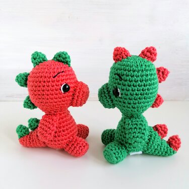 Two crocheted amigurumi dinosaurs, one red with green spikes and one green with red spikes, seated facing each other.