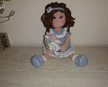 Sitzende gehäkelte Puppe mit braunen Locken, blau-weißem Kleid und kleinem buntem Einhorn im Arm.