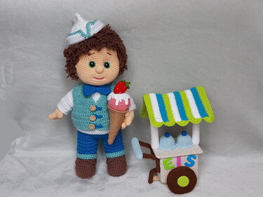 Gehäkelte Amigurumi-Puppe als Eisverkäufer mit Mütze, türkis-blauer Weste und Eishörnchen; daneben ein kleiner gestreifter Eiswagen mit Eiskugeln und Buchstaben EIS.