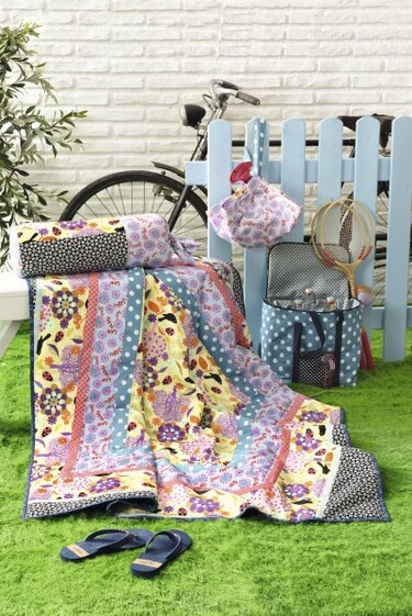 Bunte Patchworkdecke mit Blumen-, Punkte- und Marienkäfermuster, dazu passendes Kissen auf grünem Rasen vor hellem Lattenzaun und Fahrrad