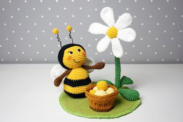 Gehäkelte gelb-schwarze Amigurumi-Biene mit weißen Flügeln neben gehäkelter Margerite und Körbchen