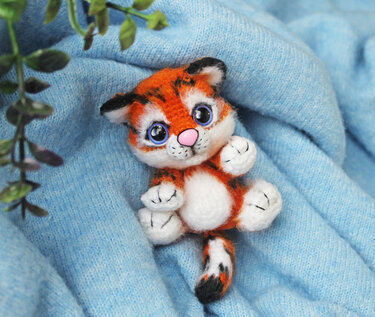 Gehäkelter Amigurumi-Tigerwelpe mit orangen Streifen und blauen Augen auf hellblauem Stoff