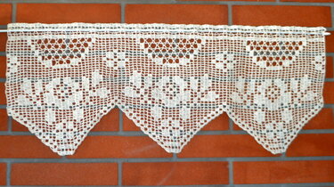 Cremefarbene gehäkelte Scheibengardine mit geometrischem Filetmuster und dreieckigem Saum vor roter Ziegelwand