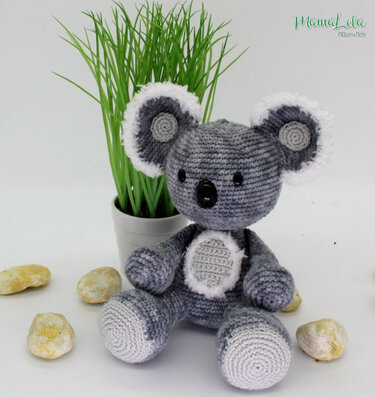 Gehäkelter Amigurumi-Koala in Grau und Weiß sitzt vor einer Topfpflanze.