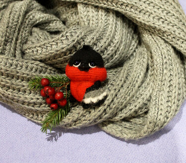 Gehäkelter Dompfaff auf grauem Strickschal mit Tannenzweig und roten Beeren