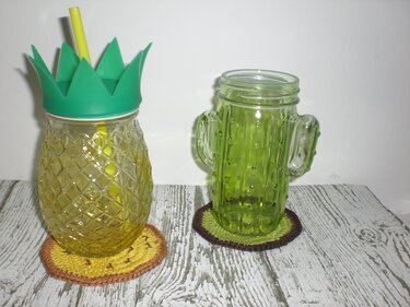 Gehäkelte runde Untersetzer unter einem Ananas- und einem Kaktusglas auf Holz