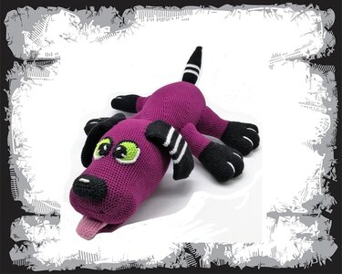 Gehäkelter Amigurumi‑Hund in Magenta und Schwarz mit grünen Augen und herausgestreckter Zunge, liegend