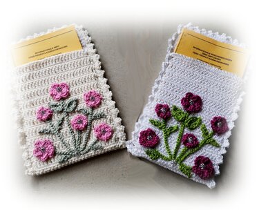 Zwei kompakte gehäkelte Hüllen mit aufgesetzten Blumenmotiven, Muschelrand und gelben Dokumenten, geeignet für Impfausweis oder kleine Heftchen