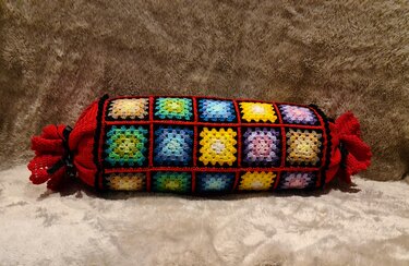 Handgehäkelter Nackenrollenbezug aus bunten Granny-Square-Quadraten mit schwarzer Einfassung und roten gerafften Enden